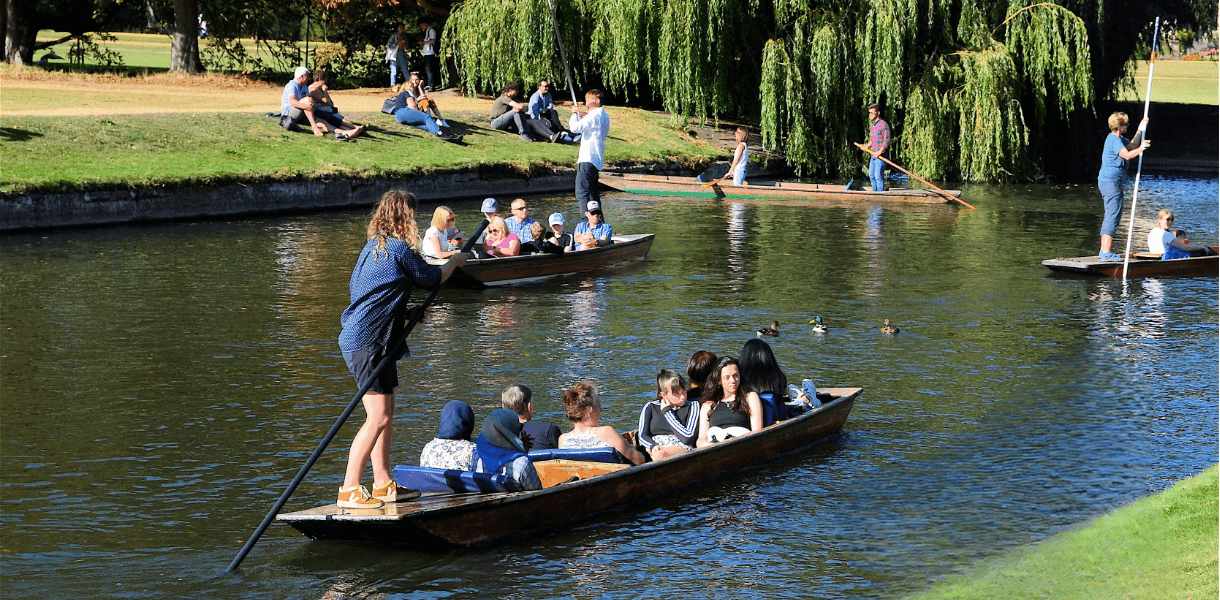People enjoying a sunny day in Cambridge, with several punts gliding along the River Cam while others relax on the grassy banks beneath willow trees.