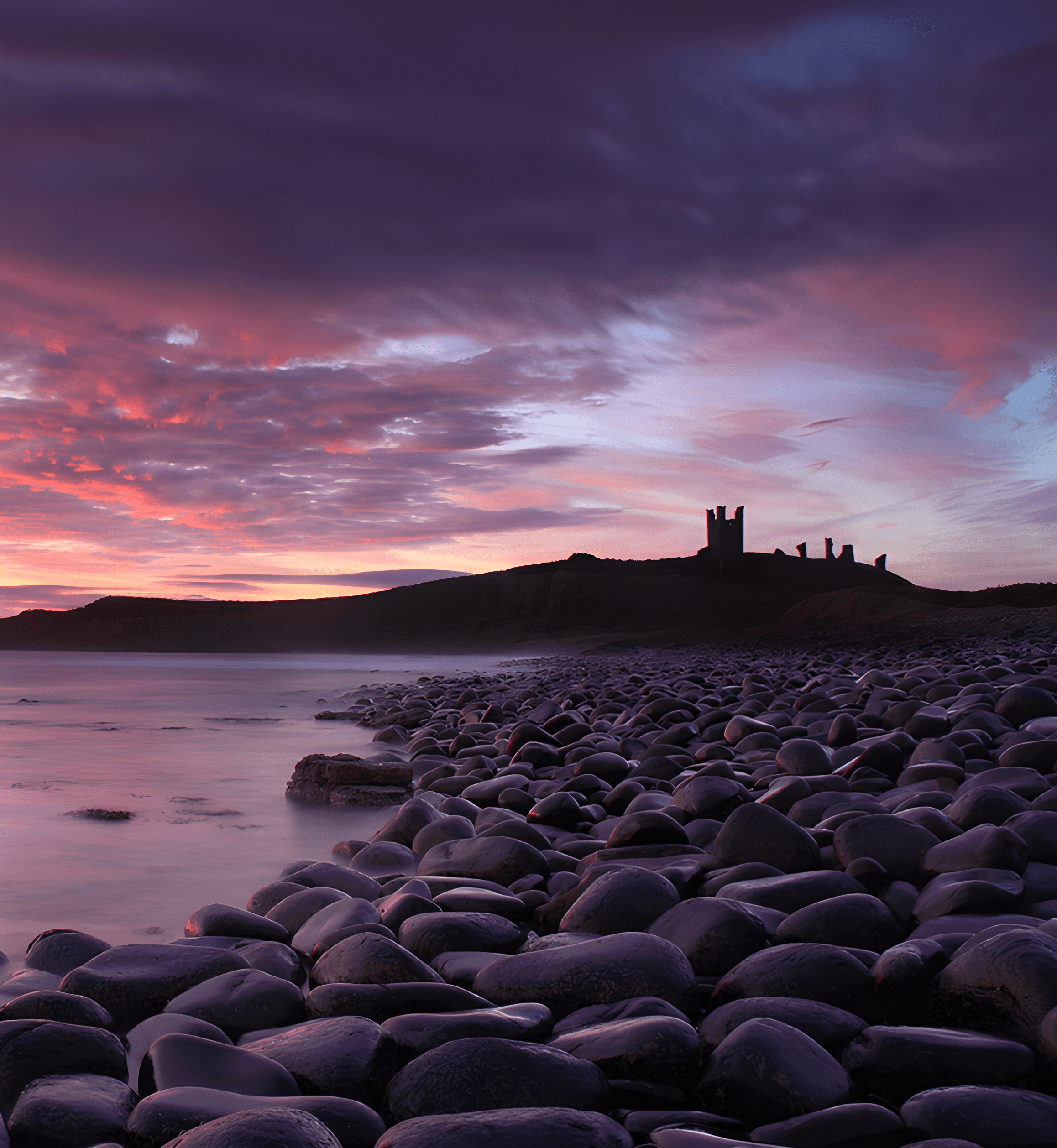 Dunstanburgh Castle, Northumberland silhouetted on a headland at dusk, seen beyond a wide beach of smooth dark stones under a dramatic pink and purple sky.