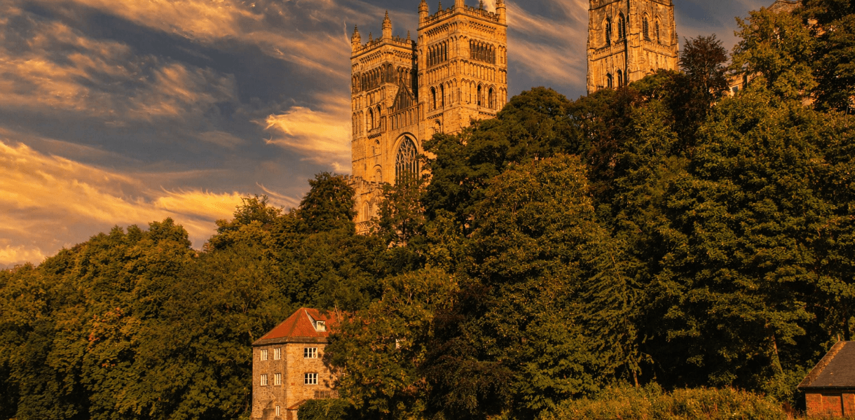Durham Cathedral rises above the wooded riverbank in warm evening light, its Norman towers glowing gold above the trees