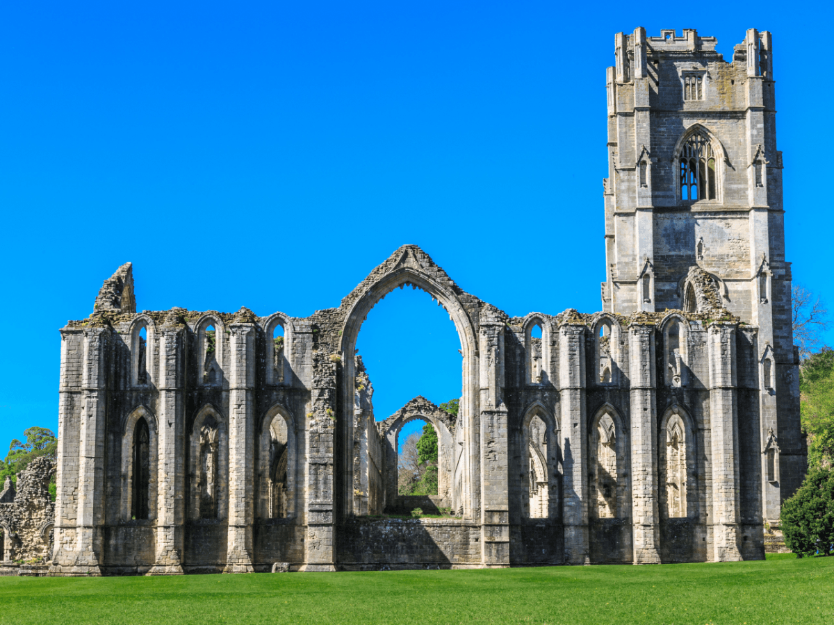 Ruins of Fountains Abbey in North Yorkshire, with tall Gothic stone arches and a square tower rising above a wide green lawn under a clear blue sky.