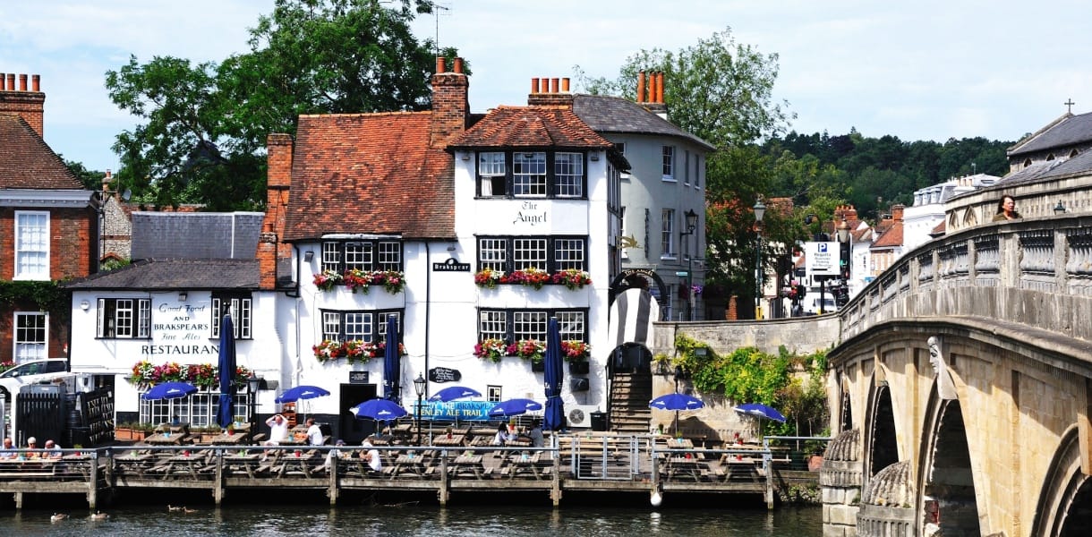 Riverside buildings and pub with outdoor seating beside the Thames in Henley-on-Thames, with flower-filled window boxes, people dining by the water, and part of Henley Bridge visible on the right.