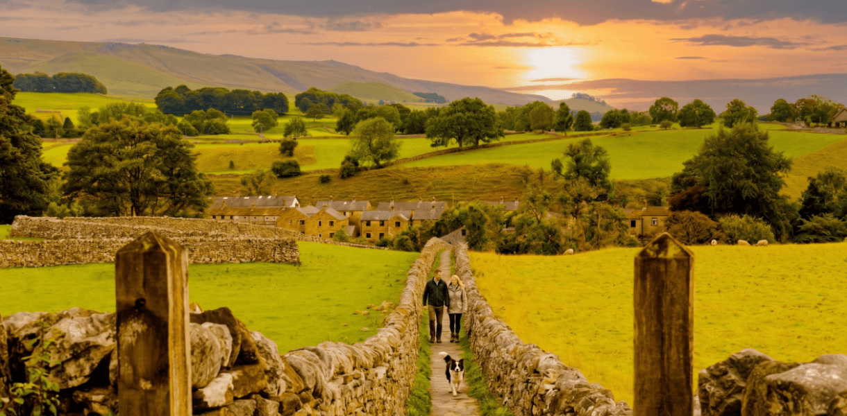 Couple and dog walking along a narrow stone-walled path through the Yorkshire Dales at sunset, with green fields, dry stone walls, scattered cottages and rolling hills stretching into the distance.