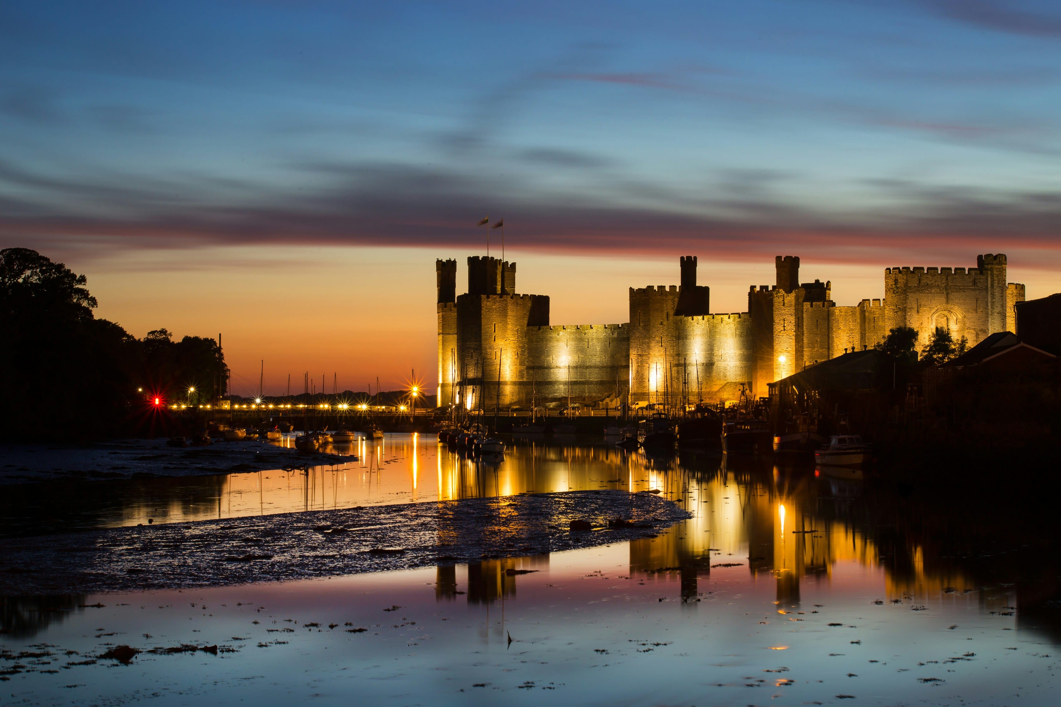 Caernarfon Castle illuminated at dusk in North Wales, reflected in the still water of the harbour, with moored boats and a deep blue and orange evening sky behind.
