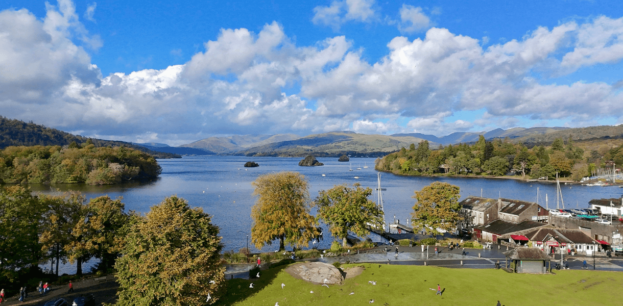 View over Lake Windermere from Bowness-on-Windermere, with calm blue water, wooded shores, small boats, autumn trees, and distant Cumbrian hills beneath dramatic clouds, illustrating an easy no-car escape to the Lake District.