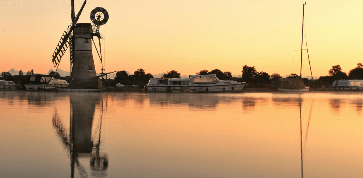 Traditional windmill and moored boats on the Norfolk Broads at sunset, reflected in still water under a soft golden sky.