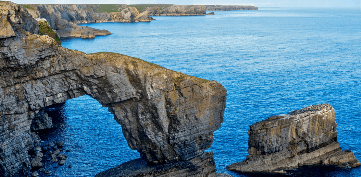 Dramatic sea arch and limestone cliffs on the Pembrokeshire coast, with blue water, distant headlands and a rocky offshore stack.