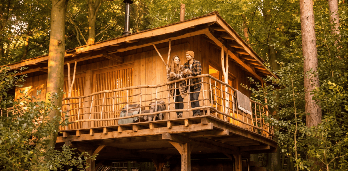 Couple standing on the balcony of a wooden woodland cabin surrounded by trees, enjoying a cosy countryside spring escape.