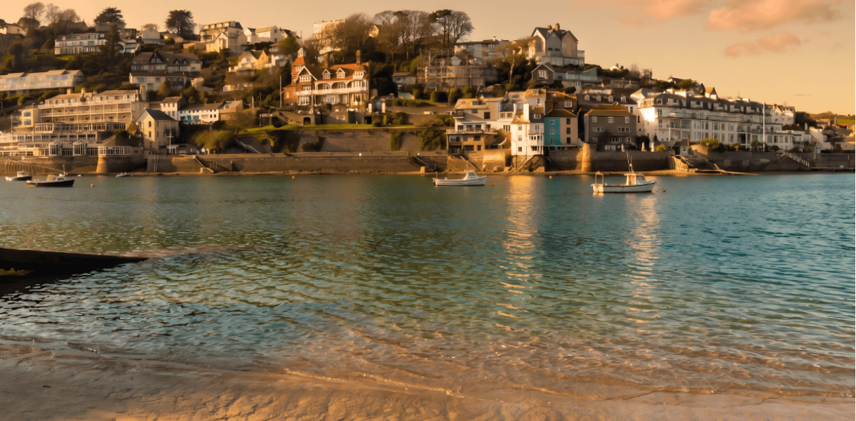 Waterfront houses and small boats in a sheltered harbour at golden hour, viewed across calm water from a sandy beach.