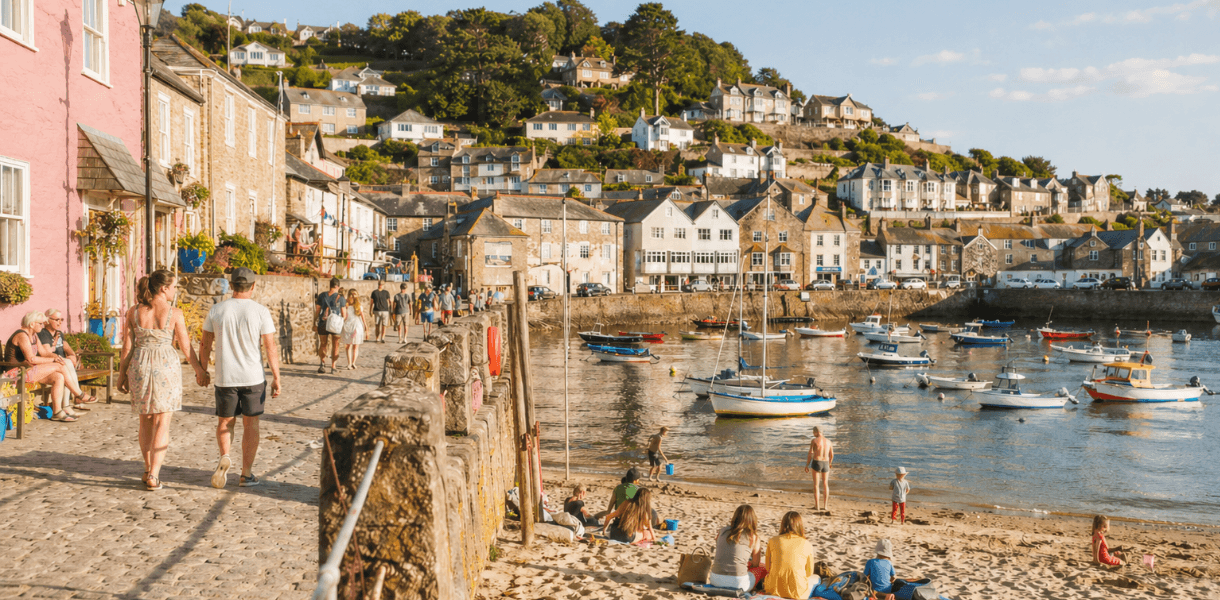 People enjoying a sunny summer day beside a harbour, with boats on the water, families on the small beach and cottages rising up the hillside.