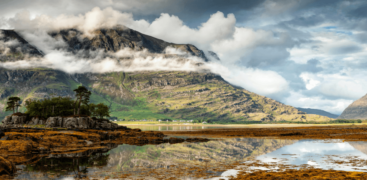 Clouds wrapping around Highland mountains above a calm loch near Torridon, with rocky shoreline and reflections in the water.