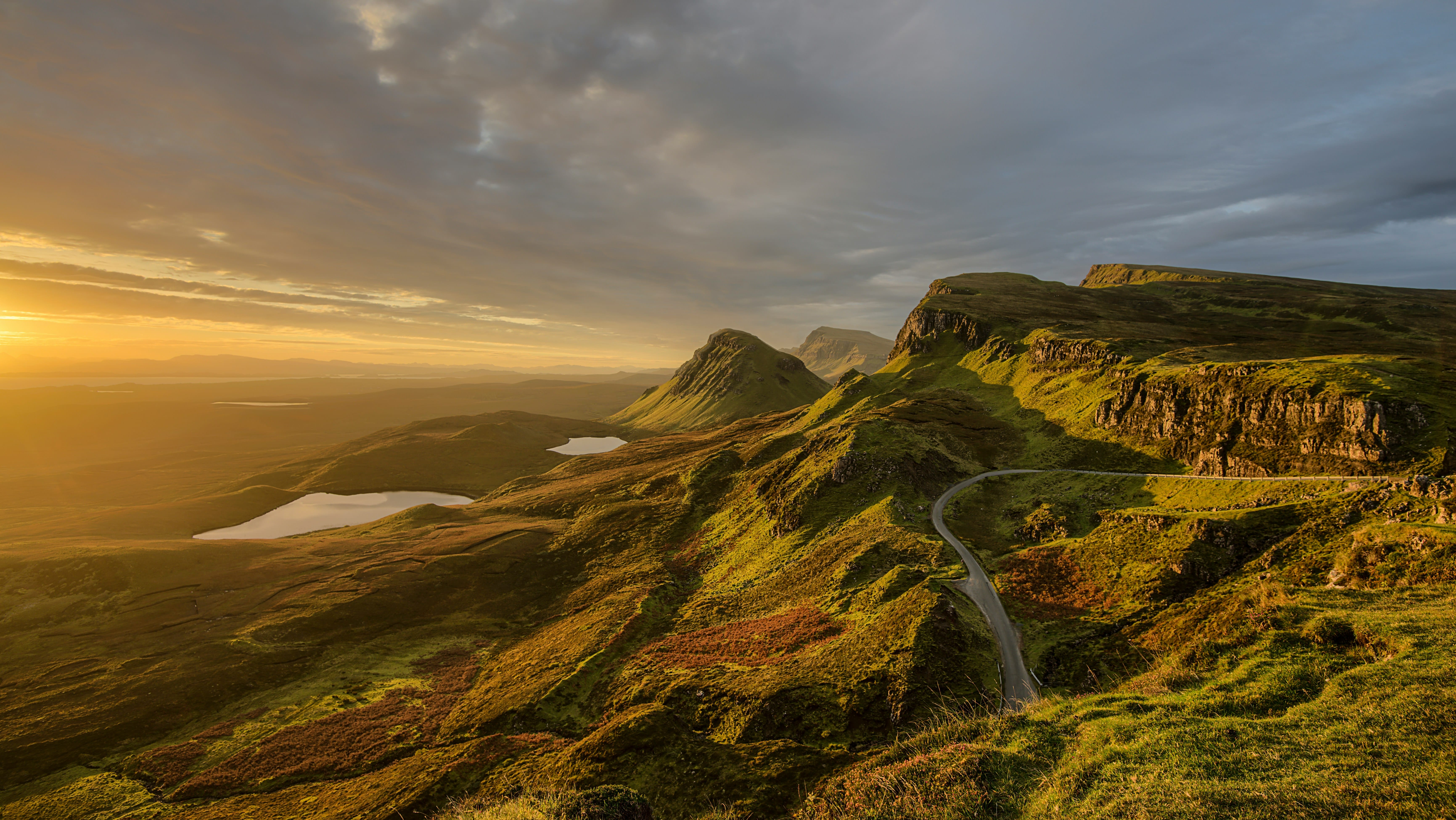 Golden sunlight over the Quiraing on the Isle of Skye, with a winding single-track road, rugged green cliffs, and small lochs stretching into the distance beneath dramatic cloud-filled skies.