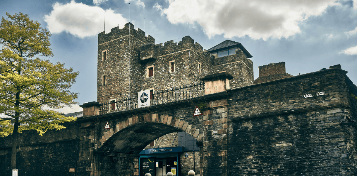 Historic city walls in Derry/Londonderry, with a stone archway, battlements and the Tower Museum entrance beneath a cloudy sky.