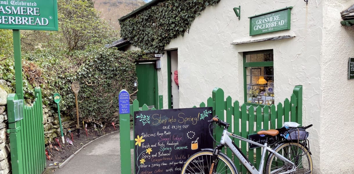 Grasmere Gingerbread shop in the Lake District, with a green picket fence, chalkboard sign and bicycle outside the whitewashed cottage building.