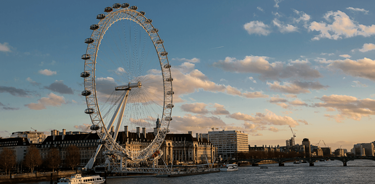 The London Eye beside the River Thames at golden hour, with Westminster Bridge, riverside buildings and boats beneath a partly cloudy sky.