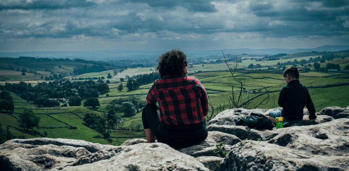 Two walkers sitting on the limestone pavement above Malham Cove, looking out across the green fields, dry stone walls and rolling hills of the Yorkshire Dales.