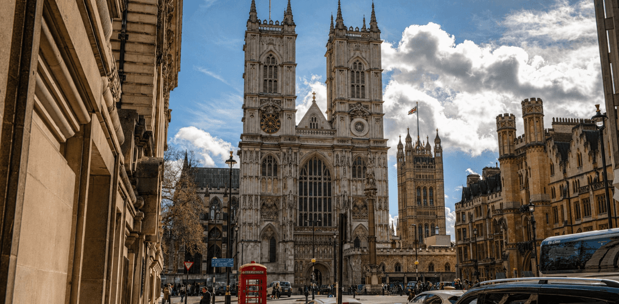 Westminster Abbey viewed from a nearby London street, with historic stone buildings, parked cars, pedestrians and a red telephone box in the foreground under a dramatic blue sky.
